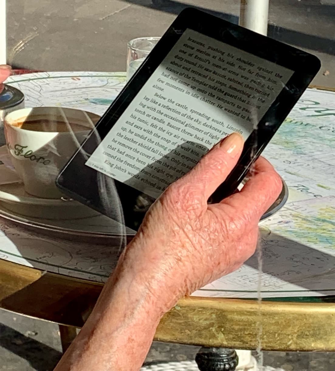 A hand holds a reader at a cafe table beside a coffee cup, the street reflected in the glass tabletop.